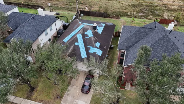 Drone inspection after a tornado in Cypress, Texas: strips and patches of blue synthetic underlayment cover every spot where the storm tore shingles off this single-story home. Patching each blown-off area individually seals the exposed decking quickly without disturbing the shingles that are still holding. Downed branches in the front yard and debris scattered across the open field behind the homes show the tornado's path through the neighborhood.