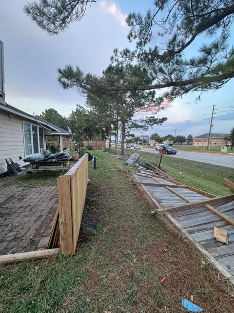 The tornado's path across the property in Cypress: whole sections of wooden privacy fence lie flat along the road, a basketball goal is knocked over, and branches and debris are scattered through the yard. Damage outside the house, like fences and trees, is documented alongside the roof and chimney so the homeowner's insurance claim reflects everything the storm destroyed.