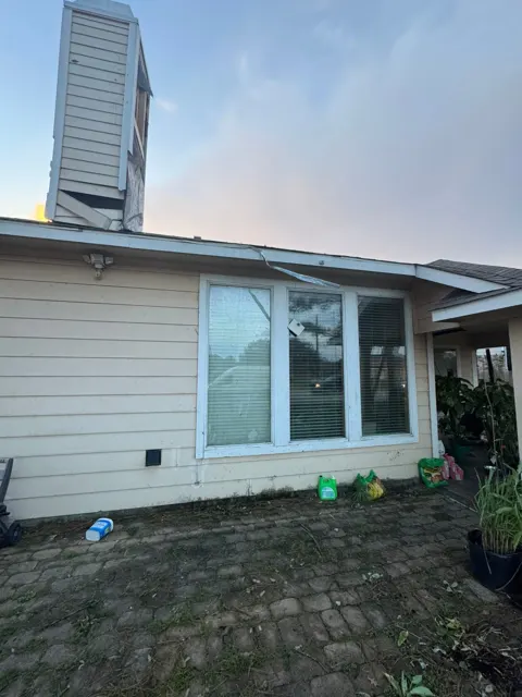 Ground view of the same home after the tornado: the chimney chase siding is peeled back and broken, and a strip of metal fascia or trim has been pulled loose from the roof edge and hangs over the front windows. Damage at the roof edge matters because once the fascia and drip edge are disturbed, wind can get under the first rows of shingles in the next storm. Photographing every damaged area from the ground and the roof builds a complete record for the insurance claim.