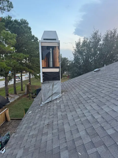 Close look at tornado damage on the roof: the siding and sheathing on one face of the chimney chase are torn away, exposing the wood framing, the metal flue pipe and tattered house wrap, with a loose piece of material draped over the shingles below. Debris is scattered across the roof. A wood-framed chimney chase like this catches wind like a sail, which is why chases are often among the first parts of a roof to fail in a tornado or severe straight-line winds.
