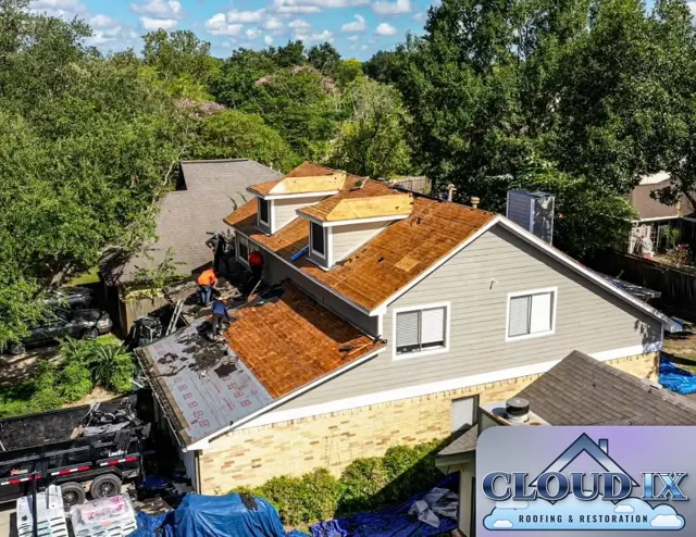 Angled drone view of the Cypress tear-off: the crew removes the last of the old shingles and felt while the two front dormers already have new OSB decking. A dump trailer in the driveway collects debris and blue tarps cover the beds and walkways around the house. Replacing damaged decking is often part of a wind and hail insurance claim when storm damage or years of leaks have weakened the wood underneath.