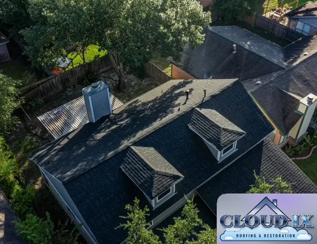 The finished roof on the Cypress home after the wind and hail insurance claim: new charcoal architectural shingles across the main roof and both dormers, with the chimney chase, plumbing vents and roof vents flashed and sealed. Dormers create extra valleys and wall-to-roof joints, so careful step flashing and shingle work there is what keeps the new roof leak-free for years.