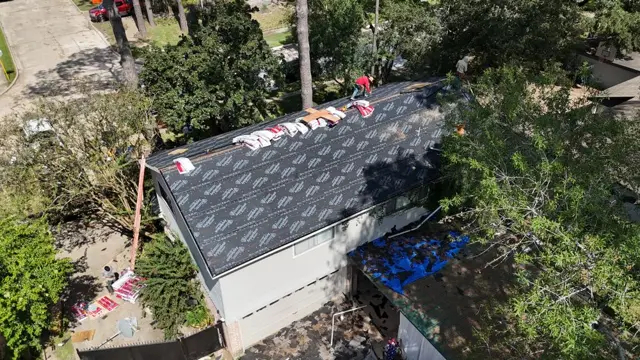 Roof replacement underway in Cypress, TX. The old shingles are torn off and the roof is dried in with dark synthetic underlayment, with new shingle bundles staged along the ridge. A tarp protects the neighboring patio roof from tear-off debris. Drying in the whole roof the same day keeps the home protected if afternoon storms roll in during the build.