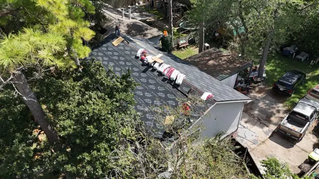 The Cloud IX crew works along the ridge on a roof surrounded by tall pines and hardwoods, with bundles staged and the first courses of new shingles going on. Working under heavy tree cover means extra care with debris, ladder placement and cleanup, all part of a cash roof replacement done right.