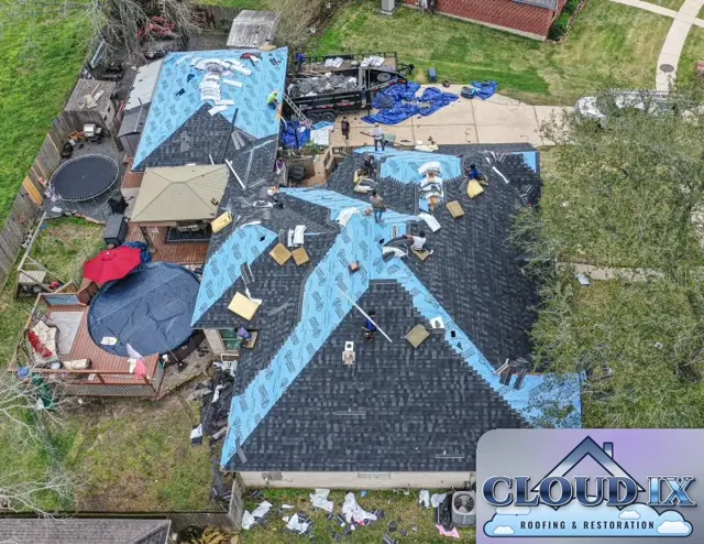 Drone view of a hail claim roof replacement in progress in Magnolia, Texas. Blue synthetic underlayment covers the hips and valleys of the complex single-story roof while the crew lays new Class 3 shingles across the field, working toward the ridges. A dump trailer and tarps in the driveway catch debris, and the backyard deck, gazebo and pool are covered and kept clear of the work. Underlayment goes down first in the valleys, where water concentrates and most roof leaks begin.