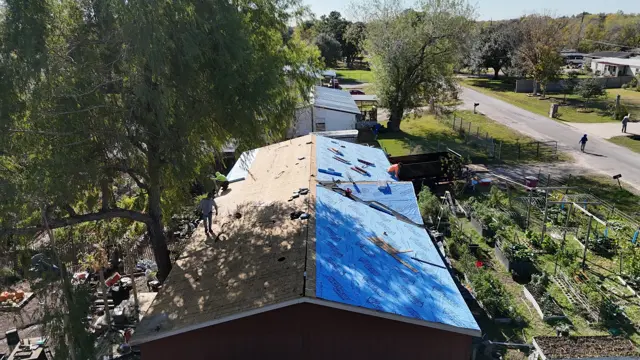 Aerial view looking down the roofline: bare decking on one slope while the other is already dried in with underlayment. Tearing off and drying in a roof in stages means the home is never left fully exposed if weather moves in, a practical concern during Houston-area storm season. The drone shot also shows crew members working safely along the ridge.