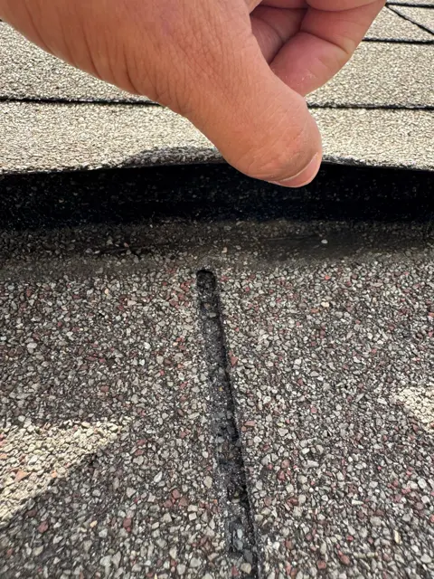 Close-up of a creased asphalt shingle with a broken seal strip and loose granules. Creasing happens when strong wind folds a shingle tab back over itself; the bend line permanently weakens the shingle even after it lies flat again, so creased tabs are treated as damaged rather than repairable. Documenting creases, broken seals and granule loss together helps establish the full scope of storm damage for an insurance claim in Pearland, Texas.