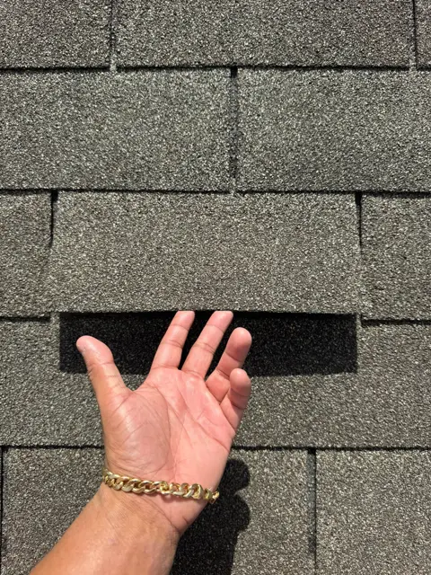 Wind damage found during a free roof inspection in Pearland, TX: a hand slides under an asphalt shingle tab that high winds pulled loose from its adhesive seal strip. Shingles are designed to bond to the course below them; once that seal breaks, the tab can lift and flap in every storm, letting wind-driven rain reach the underlayment and decking. Unsealed tabs are a key piece of evidence insurance adjusters look for when evaluating a wind damage claim.