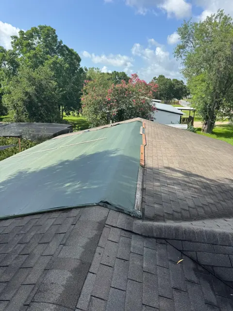 Emergency roof tarp on a storm-damaged home in Pearland, Texas. Cloud IX covered the wind-damaged slope with a heavy green tarp, fastened along the ridge cap and edges so it holds through the next rain. The opposite slope still shows the original asphalt shingles for comparison. Tarping right after a storm stops water from reaching the decking, insulation and drywall, and most homeowner policies expect you to take reasonable steps like this to prevent further damage while the insurance claim is filed.