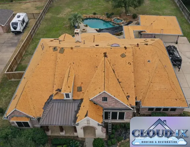 Aerial view of a full roof tear-off down to the decking on a hail-damaged brick home in Tomball, Texas, approved through the homeowner's insurance claim. Every old shingle and underlayment layer has been removed, exposing the OSB roof deck across all the hips, valleys, dormers and the detached garage roof so it can be inspected and repaired before anything new goes on. A dump trailer sits in the driveway for debris, and the pool area is covered to protect it during tear-off. Starting from the decking up means problems like soft wood or old patches are found and fixed instead of being buried under a new layer of shingles.