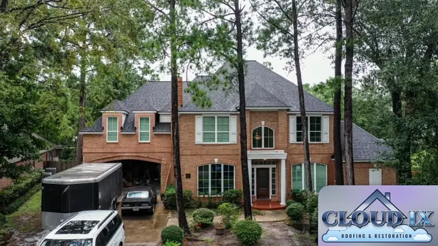 The finished roof on the Spring home seen from the street, framed by tall pines. The new charcoal shingles give the red brick two-story a clean, uniform look across the main roof and dormers. Homes under heavy pine cover get constant needle and limb debris, so a well-installed, impact-rated roof with tight ridge and valley details holds up better between storms.