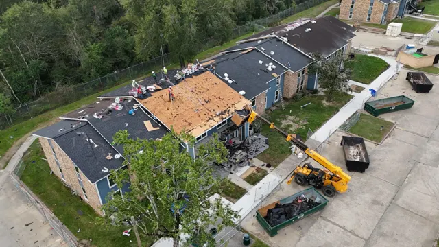 Angled aerial view of the apartment building mid-tear-off: a crew in hard hats and harnesses works on bare decking while the telehandler's debris box sits at the roof edge to catch the old shingles. Tarps cover the ground and walkways below, and fall-protection ropes run across the roof from the ridge anchors. Removing debris by machine instead of throwing it to the ground keeps stairways, balconies and landscaping clear on an occupied property.