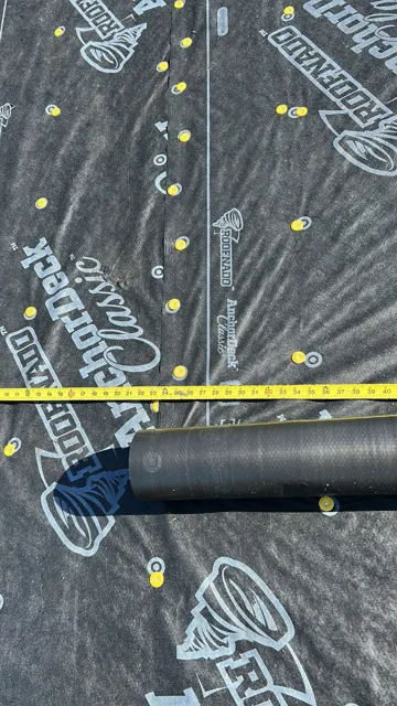 Close-up of the synthetic underlayment installed over the decking on the multi-family project, fastened with plastic cap nails in a tight, even pattern and measured with a tape to confirm proper laps at the seams. Cap nails hold synthetic underlayment flat and resist tearing in wind far better than staples, and correct overlap is what keeps water out if a shingle is ever damaged. A roll of underlayment sits ready for the next course.