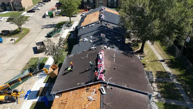Drone view looking down the length of the apartment building, with every stage of the reroof visible at once: bare decking in the foreground, synthetic underlayment, new shingles and bundles staged on the ridges. The telehandler unloads debris into a roll-off on the left while the crew starts shingling the next section. Working in planned stages lets each building be dried in the same day it is opened up.