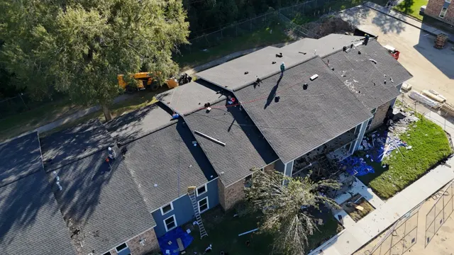 The new architectural shingles nearly finished across the step-down roof sections, with the crew working final ridge and transition details. The telehandler waits beside the building for the last round of debris. On multi-family properties, finishing building by building means each set of residents gets a sealed roof as quickly as possible.