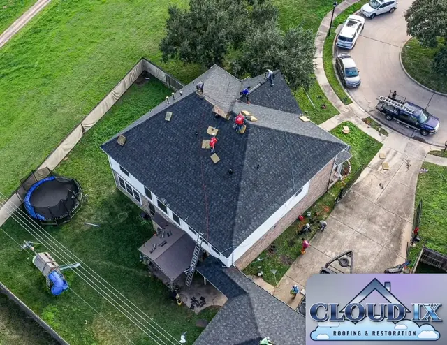 Drone view of a reroof in progress on a two-story brick home in Tomball, Texas. The Cloud IX crew works across the hips and ridge of the main roof while new shingle bundles sit staged on the slopes and the work truck waits at the curb. A privacy fence screen lines the backyard, and the yard below is kept clear so cleanup goes quickly when the roof is finished.