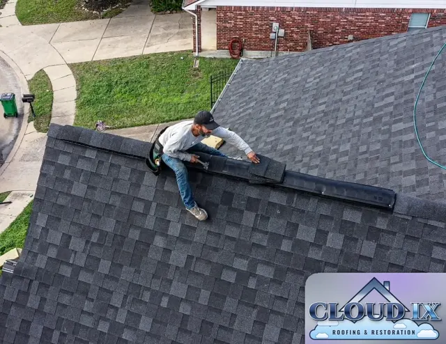 An installer nails cap shingles over a continuous ridge vent along the peak of the roof. Ridge vents let hot, moist air escape from the attic along the full length of the ridge, which helps lower attic temperatures through a Texas summer and protects the roof deck and shingles from heat and moisture buildup. Each cap shingle overlaps the last so the vent stays covered and watertight.
